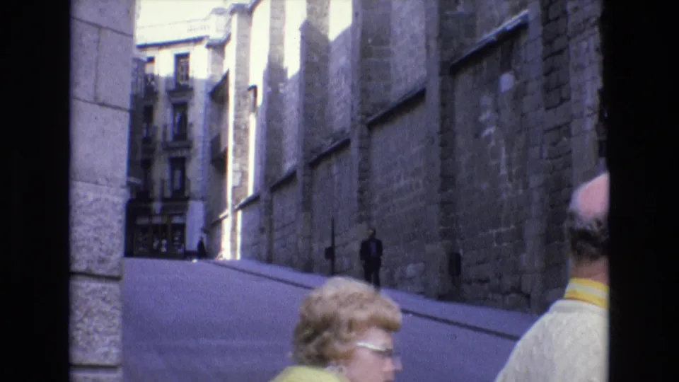 Spain 1972 frame looking down a narrow street between stone buildings, with a blonde woman wearing glasses and another pedestrian partly visible in the foreground.