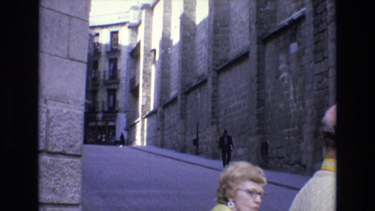 1972 Spain street view with tall stone architecture, pedestrians on a sloped roadway, and a woman in glasses and a yellow jacket at the lower foreground.