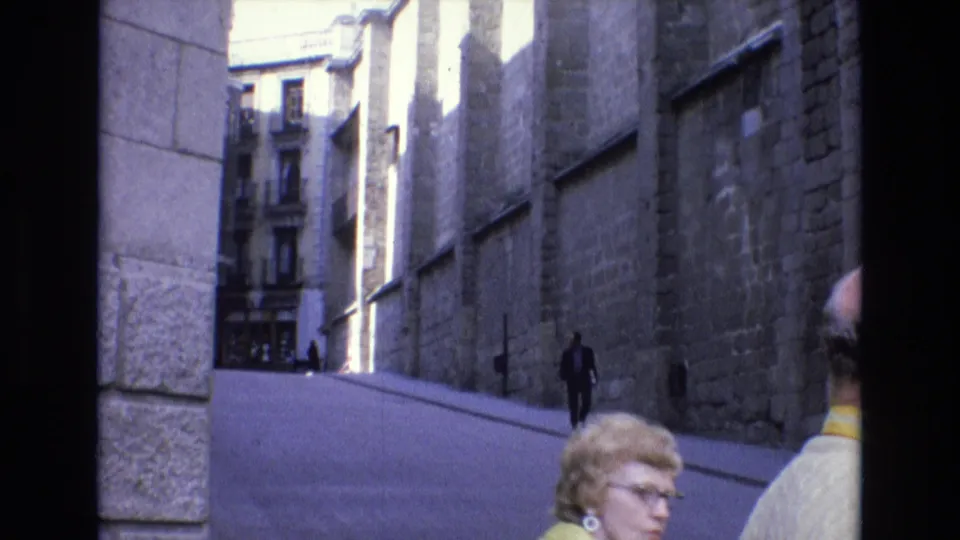 1972 Spain street view with tall stone architecture, pedestrians on a sloped roadway, and a woman in glasses and a yellow jacket at the lower foreground.