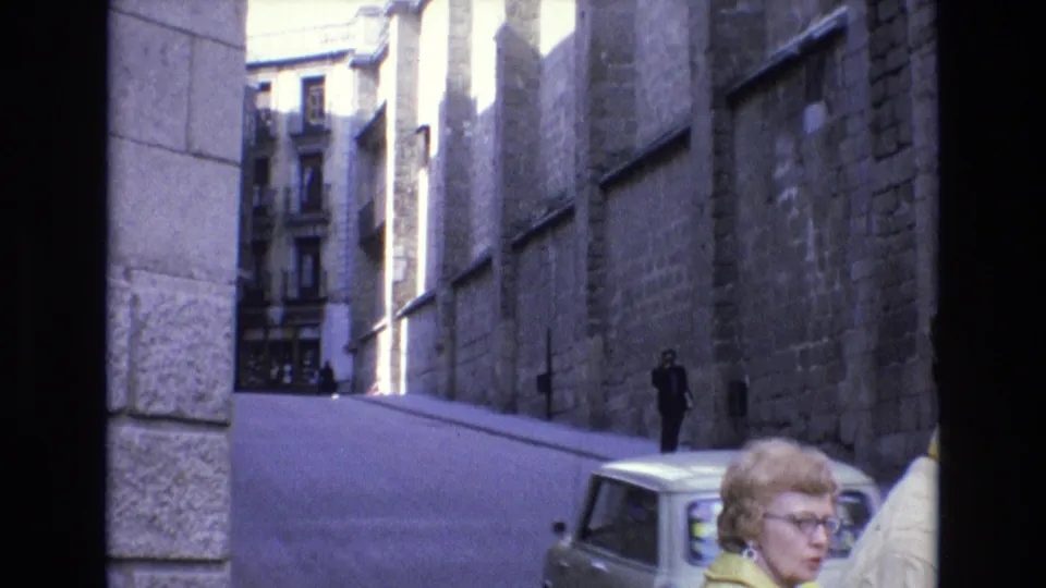 A light-colored compact car drives through a narrow Spanish street in 1972, with stone walls, pedestrians, and the woman in a yellow jacket partly visible in the foreground.