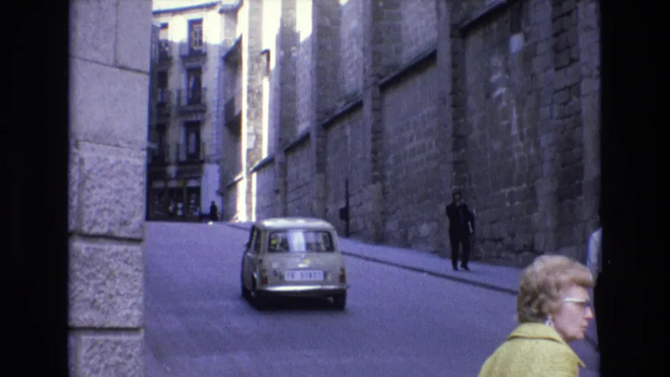 The rear of a light-colored compact car moves along a sloped street in Spain, framed by stone walls, pedestrians, and a foreground woman in a yellow jacket.
