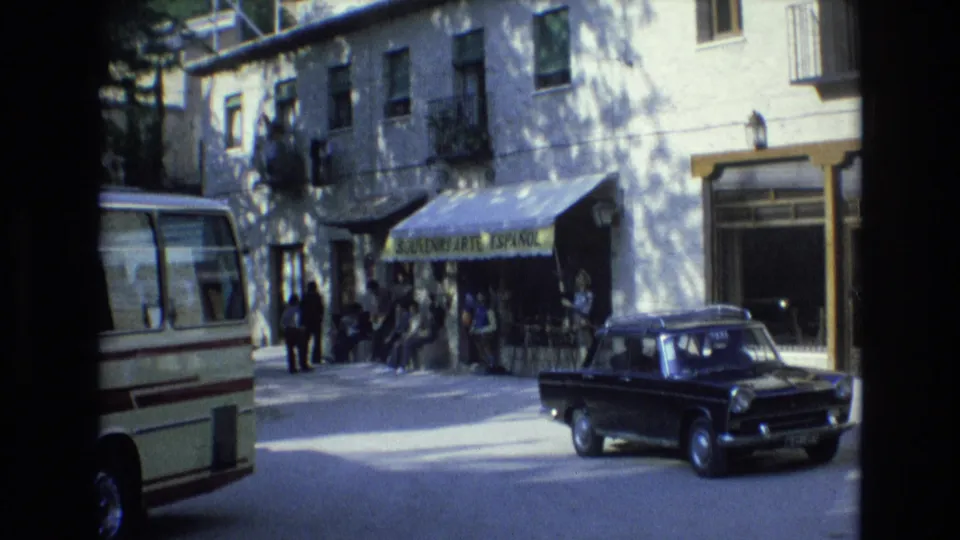 A Spanish street corner in 1972 shows a white bus, black sedan, pedestrians, and a storefront awning with Spanish souvenir-related signage.