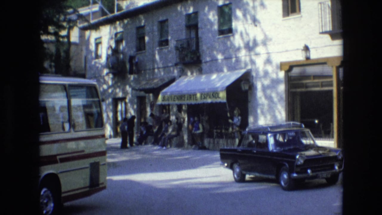 Sunlit street with a cream minibus, black sedan, and people near a souvenir shop awning.