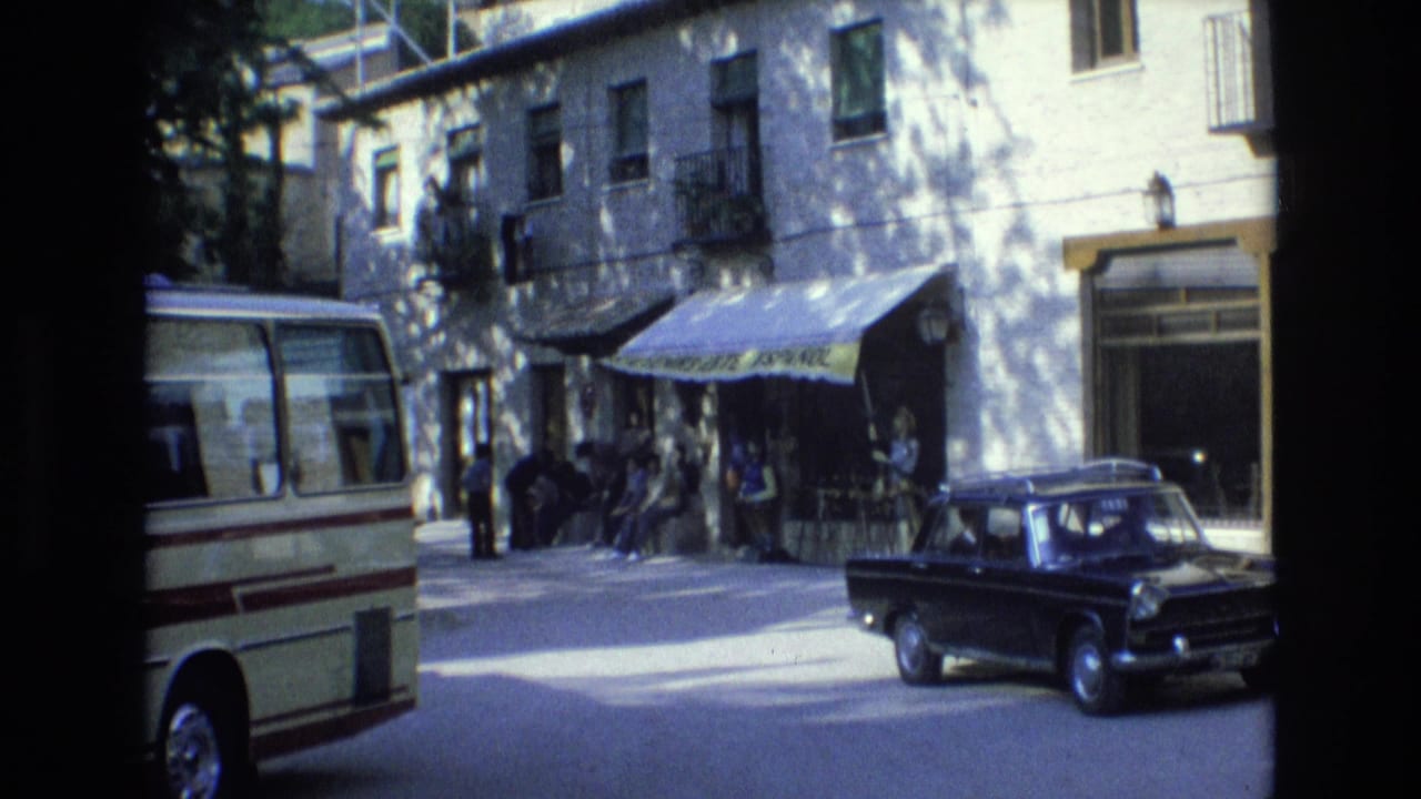 Black sedan passing a souvenir shop where people sit and stand under a yellow awning.