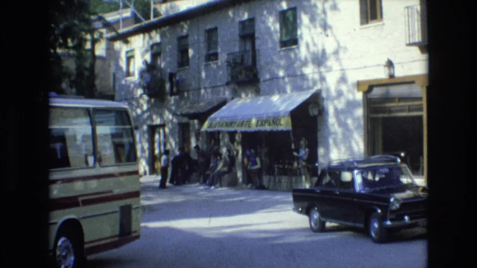 1972 Spain street view with the souvenir shop awning centered, pedestrians at the storefront, a cream minibus along the curb, and a black sedan near the right edge.