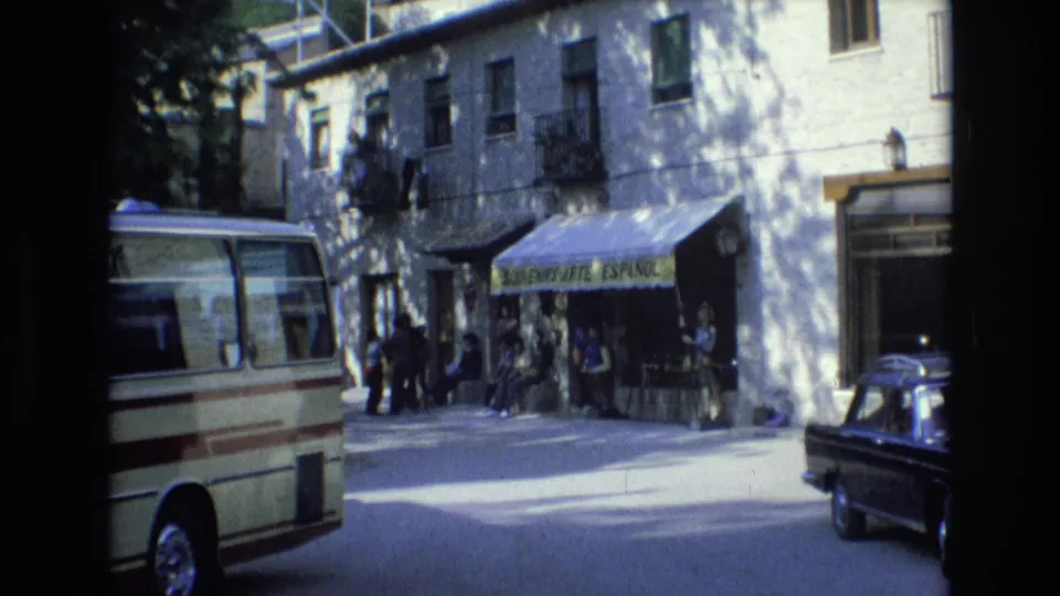 1972 Spain home-movie frame showing a cream minibus at left, people near a yellow “Souvenirs Arte Español” awning, and the front edge of a black car leaving the frame.