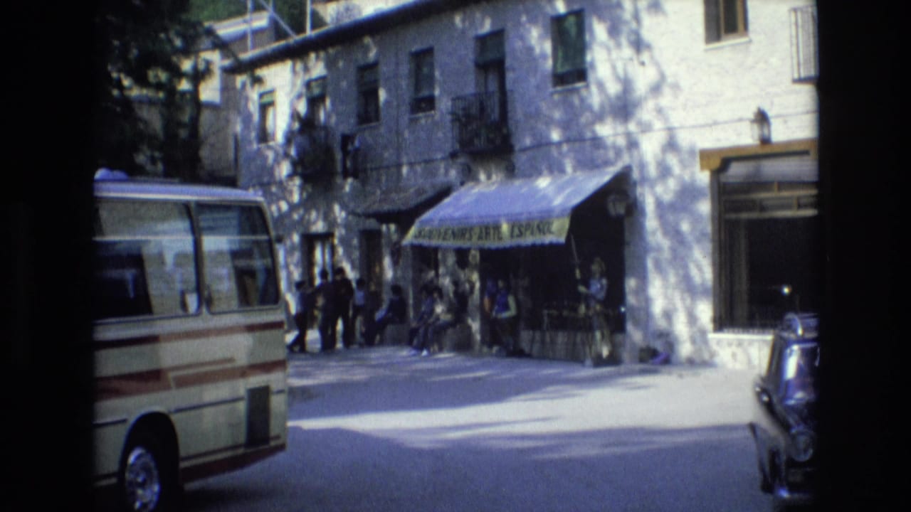 1972 Spain 8mm frame showing a cream minibus parked beside people gathered outside a storefront with a yellow souvenir-shop awning.