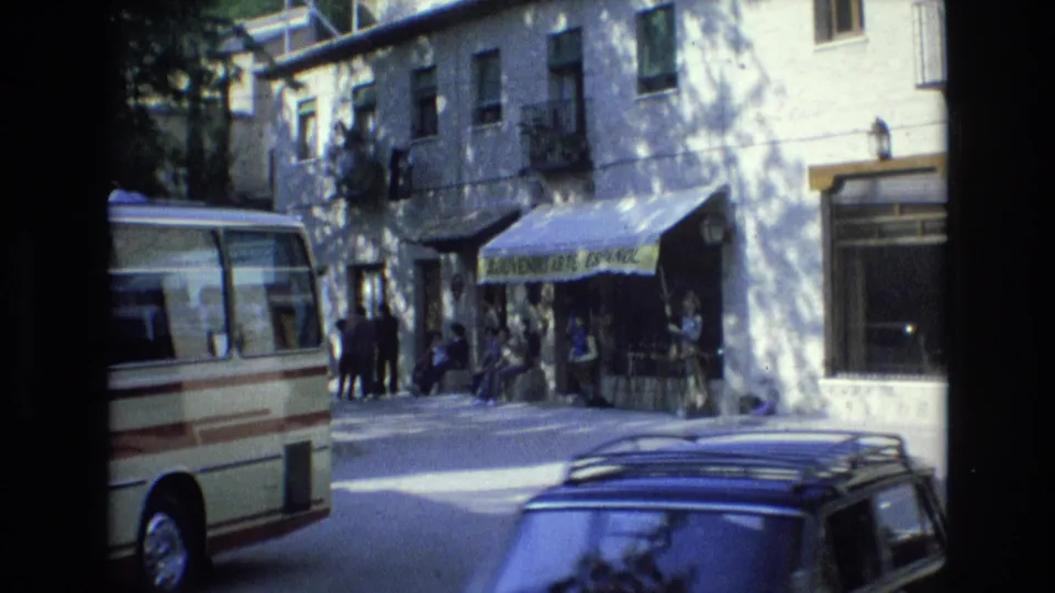 1972 Spain street view with a cream minibus, pedestrians near the souvenir-shop awning, and a dark sedan roof rack crossing the foreground.