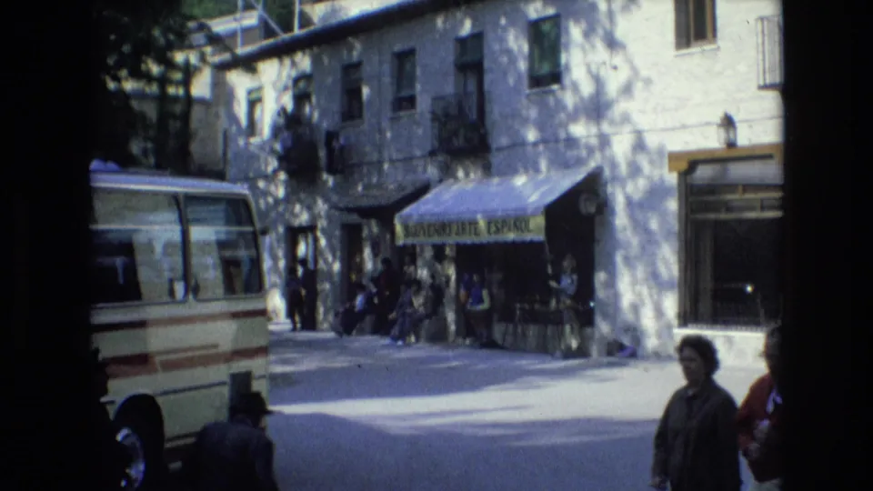 1972 Spain home-movie frame of people gathered outside a storefront beneath a yellow awning, with a parked cream minibus at the curb.