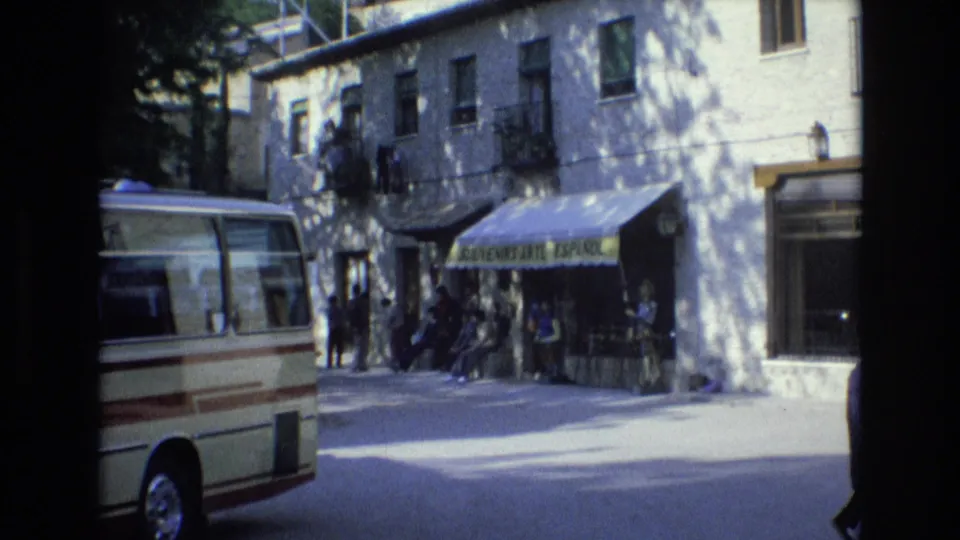 1972 Spain street frame showing the souvenir-shop awning, people gathered beneath it, a parked cream minibus, and sunlit pavement.