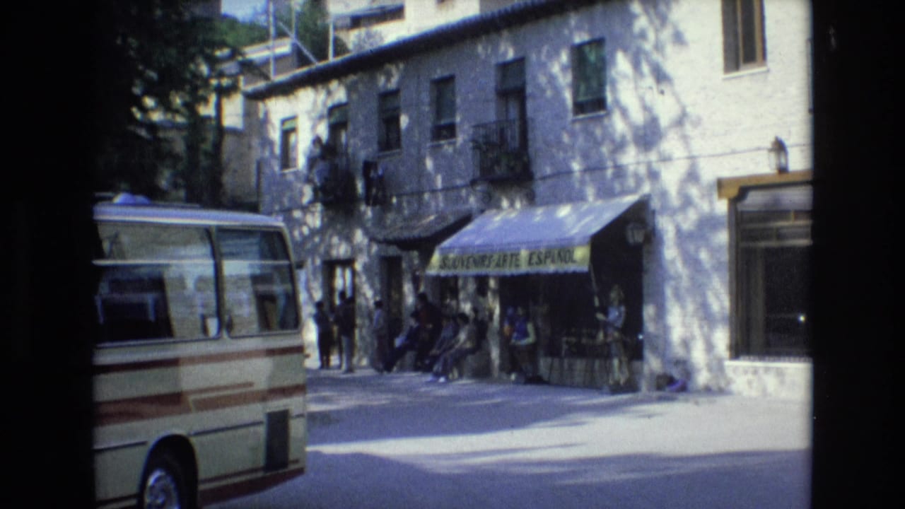 1972 Spain 8mm frame showing a cream and brown minibus near a white building, a green souvenir shop awning, and several people gathered along the sidewalk.