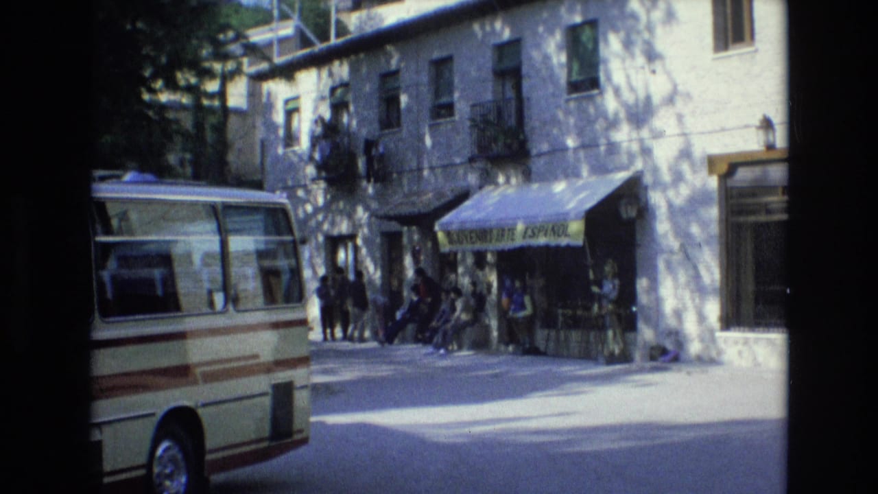 Street view of a minibus, shop awning, and pedestrians outside a white building.