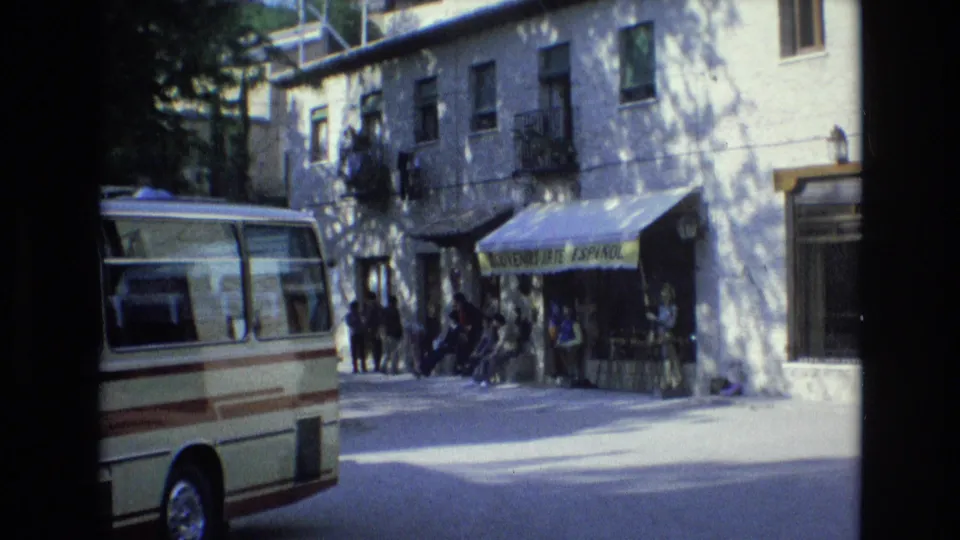 1972 Spain street frame with the minibus partly in view, pedestrians near a green awning reading as a souvenir shop sign, and balconies above the storefront.