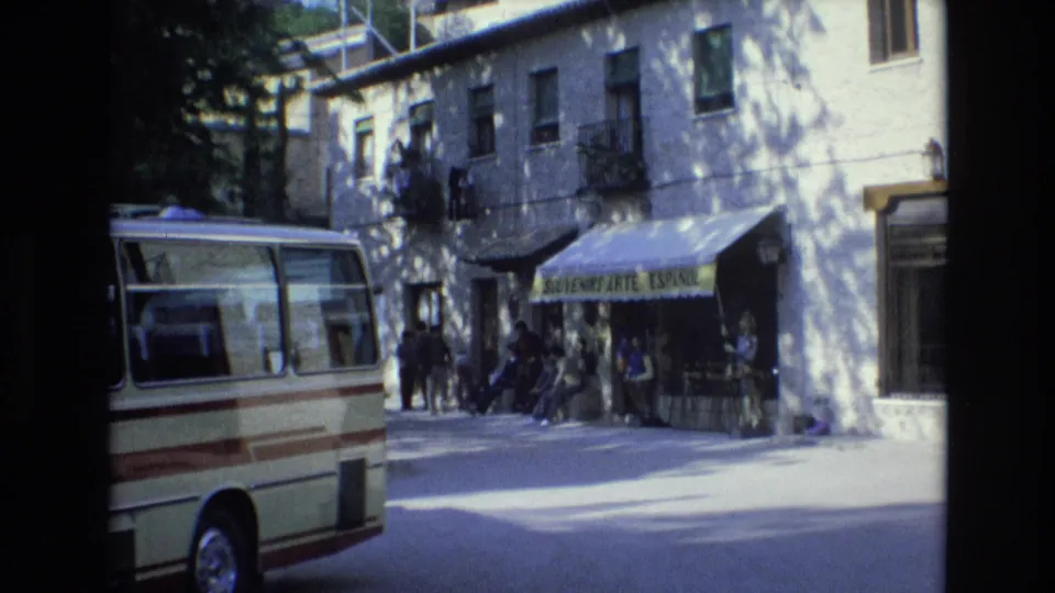 1972 Spain home-movie frame showing a parked cream and brown minibus, a sunlit storefront with a green souvenir awning, and people standing or sitting outside.