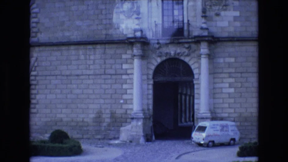 1972 Spain 8mm frame showing a formal stone facade, an ornate arched entrance with columns, a balcony above, trimmed greenery, and a small white van near the gateway.