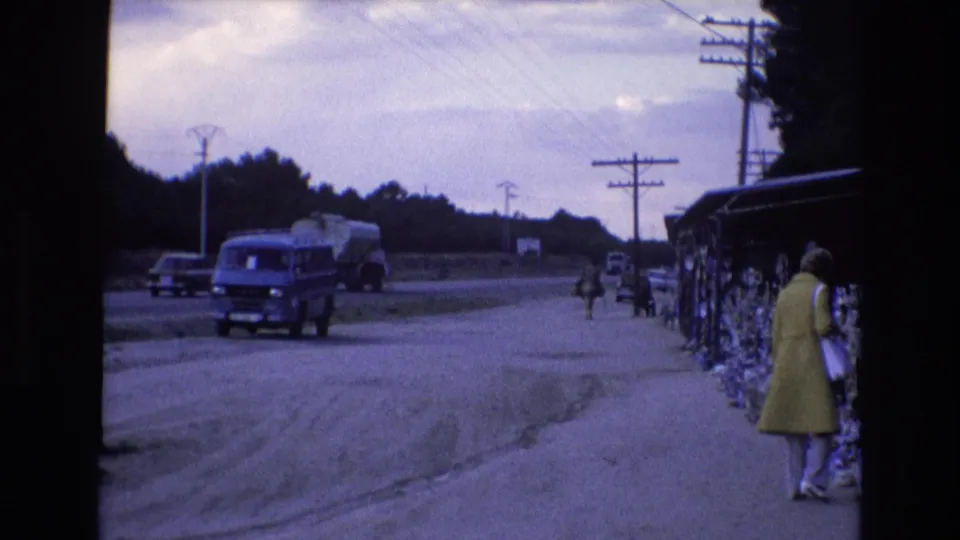 1972 Spain home-movie frame showing a blue van on the highway shoulder, roadside goods displayed under a stall, and a woman in a yellow coat at the edge of the market.