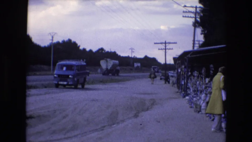 Late frame of the Spain 1972 roadside stop shows a blue van near the foreground, a cement mixer truck farther along the road, and a woman browsing beside the vendor display.