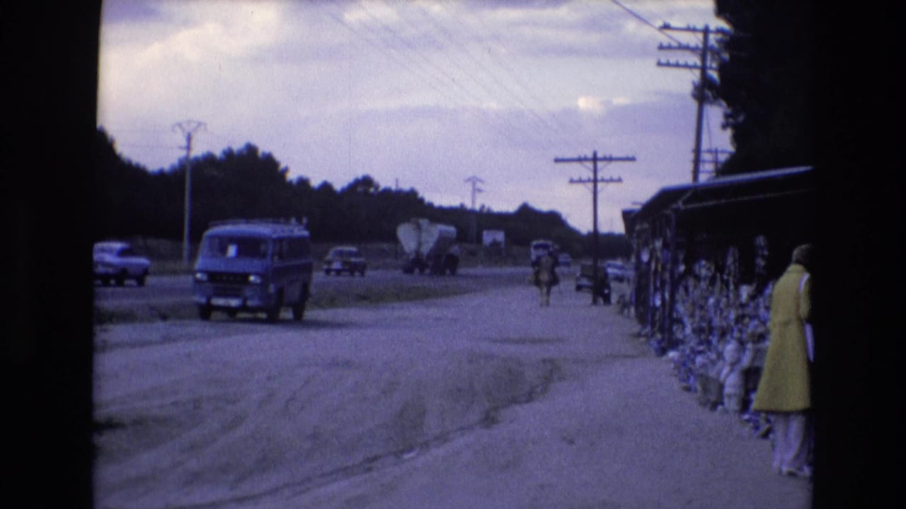 In 1972 Spain, a blue van drives along a dusty roadside while a woman in a yellow dress stands near covered sidewalk displays.