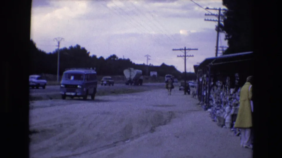 In 1972 Spain, a blue van drives along a dusty roadside while a woman in a yellow dress stands near covered sidewalk displays.