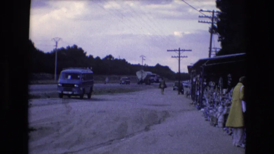 A Spanish roadside scene from 1972 shows a blue van moving past distant traffic while a woman waits near a covered market-style display.