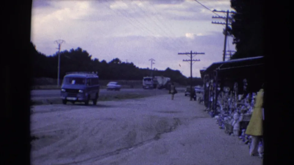 The 8mm frame shows the blue van farther along the roadside, with background vehicles, utility lines, and the yellow-dressed woman at the edge of the stall.