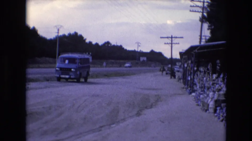 At the end of the 1972 Spain clip, the blue van continues past the roadside stall, with distant cars and utility poles receding along the road.