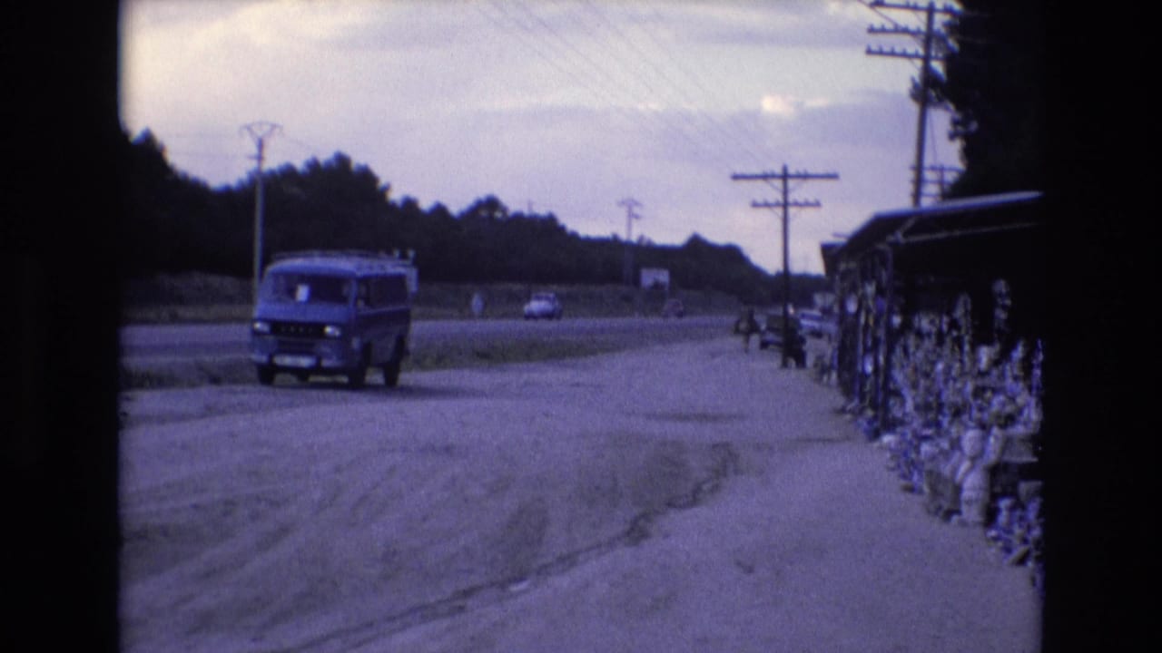 1972 Spain home-movie frame showing a blue van near the shoulder of a dusty road, with power lines, distant traffic, trees, and roadside goods at right.