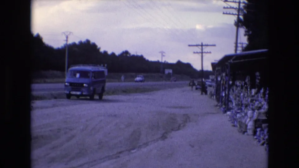 1972 Spain home-movie frame showing a blue van near the shoulder of a dusty road, with power lines, distant traffic, trees, and roadside goods at right.