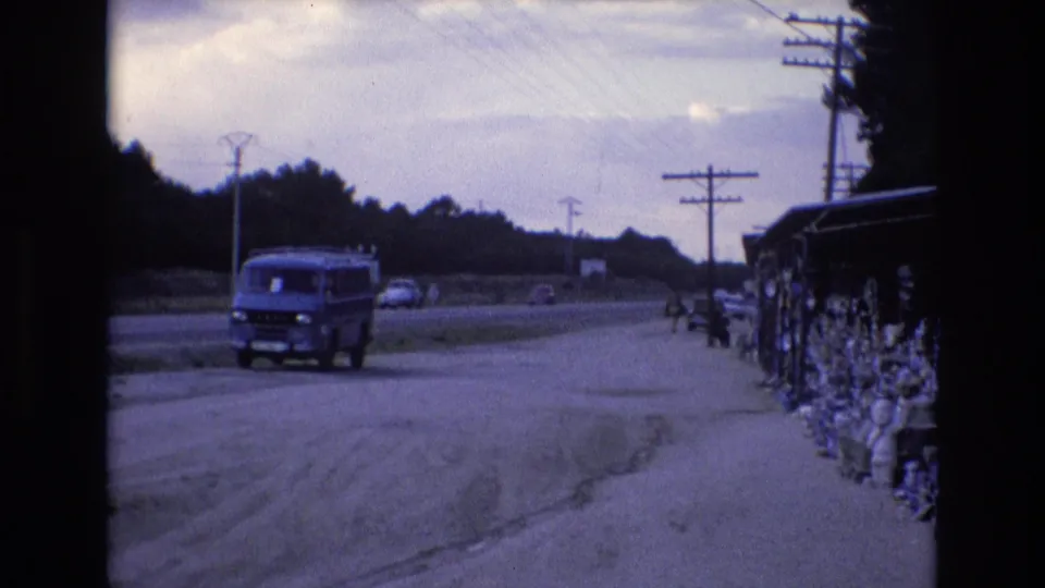 1972 Spain 8mm frame with a blue van near the road, distant vehicles on the roadway, utility poles, overhead wires, trees, and a roadside stand or display.