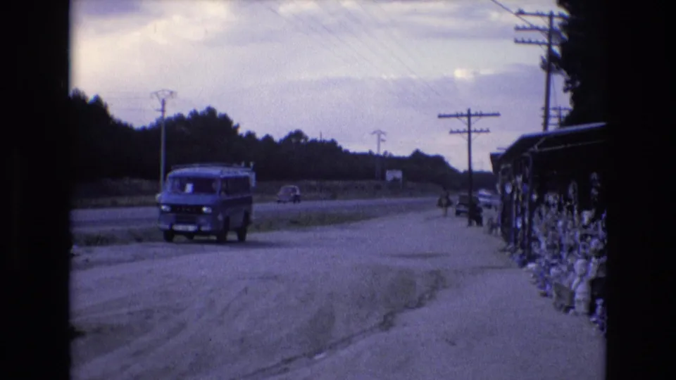 1972 Spain home-movie view of a parked blue van on a rural roadside, with a small car in the distance, power poles, a tree line, and goods displayed near the right side of frame.