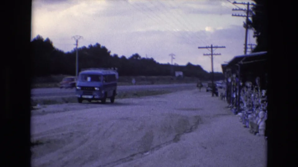 1972 Spain 8mm frame showing a blue van beside the road while a distant car passes, with utility lines, roadside goods, trees, and a cloudy sky in view.