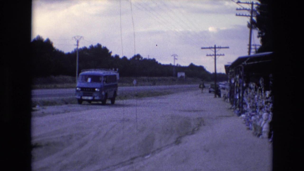 In Spain in 1972, a blue van travels along a pale roadside near utility poles, open ground, and a line of trees, seen through soft 8mm color.