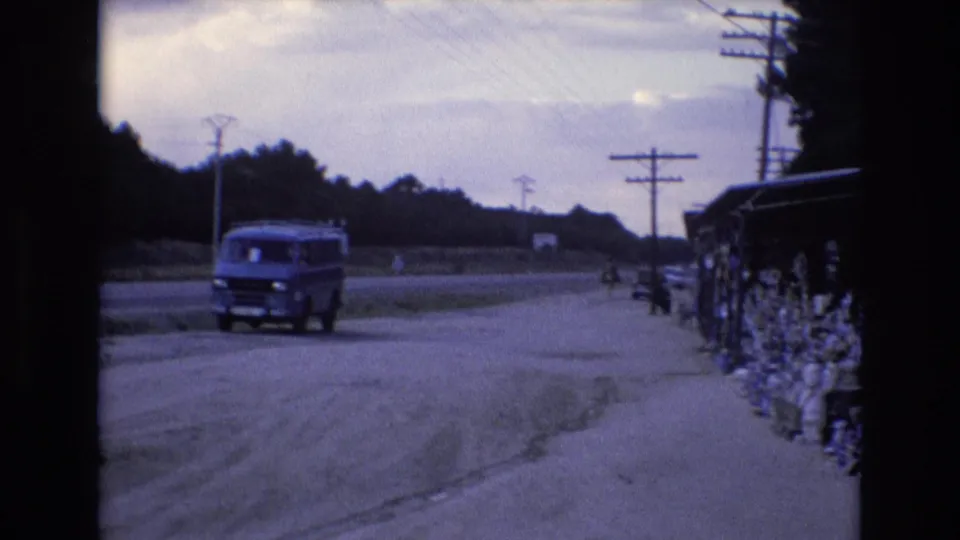 A blue van moves along the Spanish roadside in 1972, with utility poles, a shop edge, and roadside displays visible at the side of the frame.