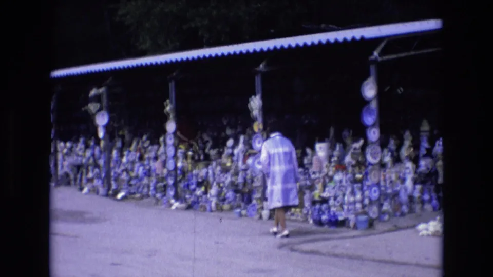 A woman in a light-colored coat approaches an outdoor roadside pottery stall in Spain in 1972, with rows of blue and white ceramics arranged under a corrugated roof.