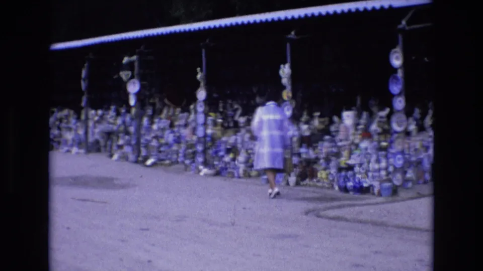 The woman continues along the roadside pottery shop in Spain in 1972, passing shelves and hanging displays of blue and white ceramic goods beneath a low roof.