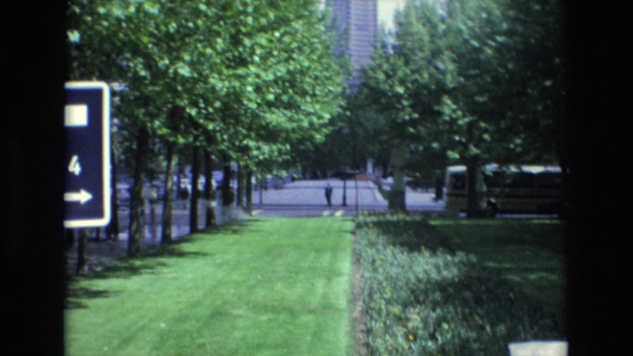 1972 Spain home-movie frame showing a green urban park edge, a blue directional sign marked 4, rows of trees, and a bus parked along the right side.