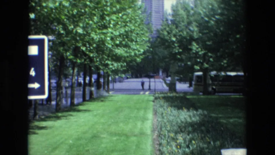 1972 Spain home-movie frame showing a green urban park edge, a blue directional sign marked 4, rows of trees, and a bus parked along the right side.