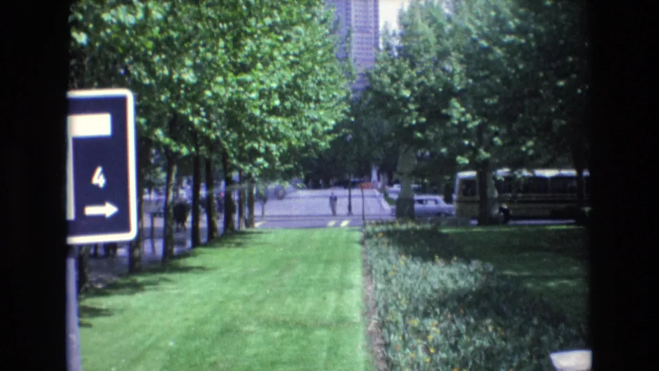 1972 Spain home-movie still showing a park-side lawn, clipped hedges, a directional sign on the left, a bus on the right, and a white car passing in the background.