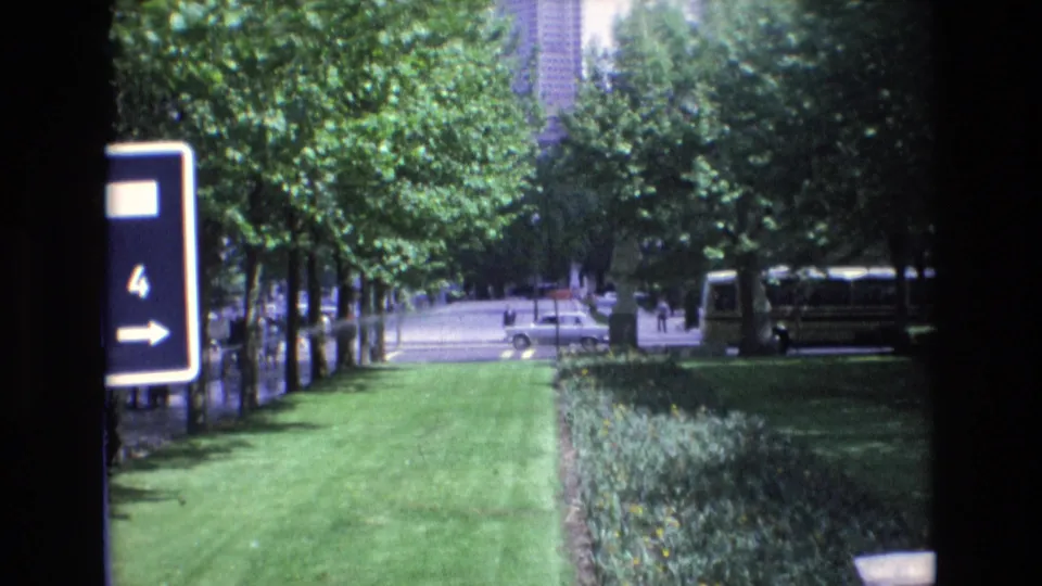 1972 Spain frame of a landscaped roadside area with a blue sign marked 4, trees casting shade, a bus to the right, a white car on the road, and distant pedestrians.