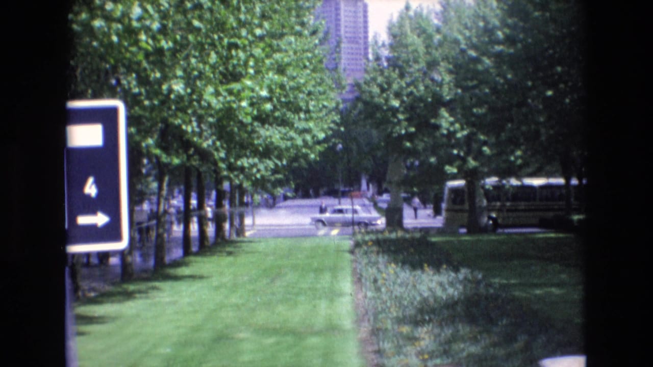 1972 Spain 8mm frame showing a green urban park edge with a directional sign, trees, a bus or coach, cars, and a tall building in the distance.