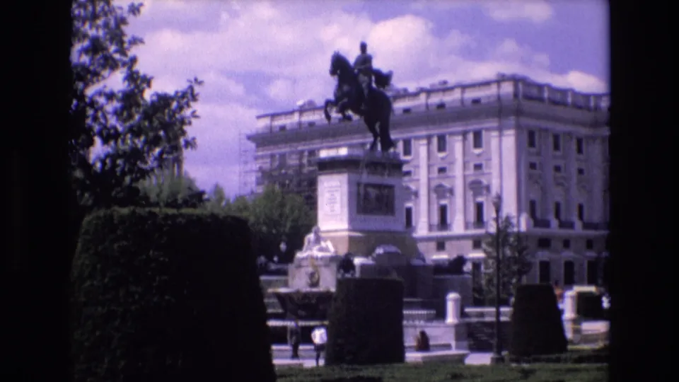 1972 Spain 8mm frame of an equestrian statue on a pedestal in a landscaped public park, with hedges, pedestrians, cars, and a pale classical building in the background.