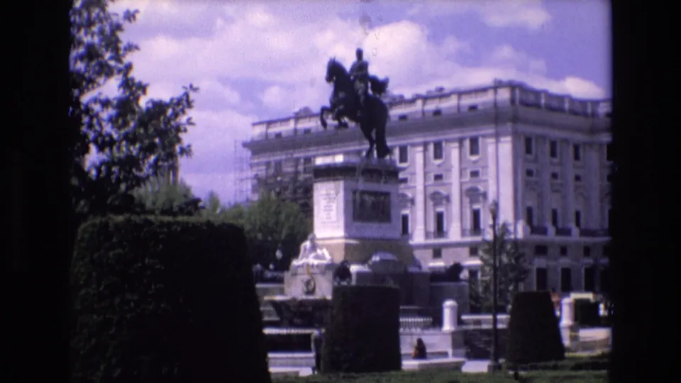 1972 Spain 8mm frame showing a closer tourist view of an equestrian monument in a formal park, framed by hedges and backed by a large light-colored public building.