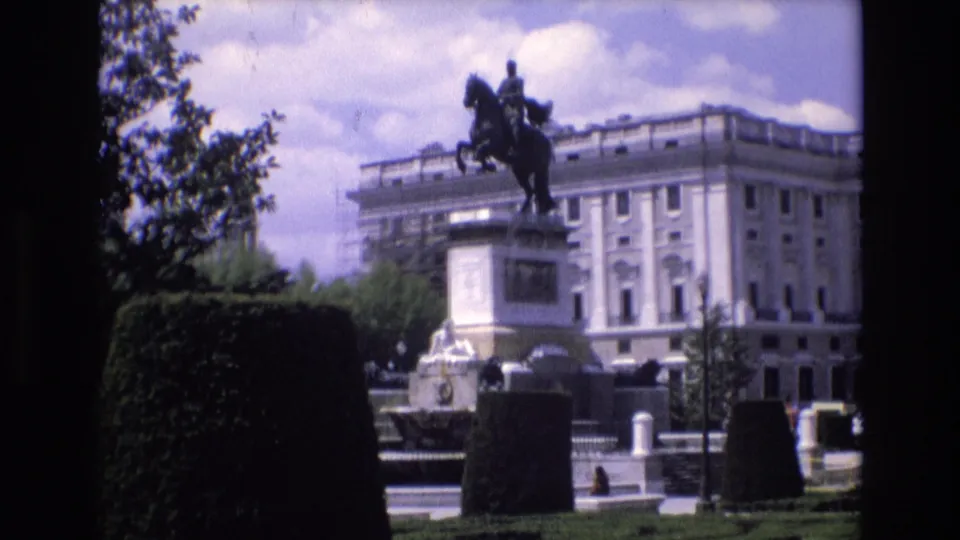 1972 Spain 8mm frame of the equestrian statue and surrounding formal park, with trimmed hedges, a light classical facade, pedestrians, and cars visible beyond the monument.
