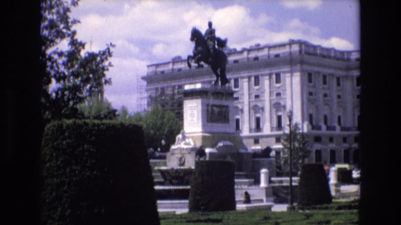 1972 Spain 8mm frame showing the Monument to Philip IV in Plaza de Oriente from across trimmed hedges, with a palatial facade and a few pedestrians in the background.