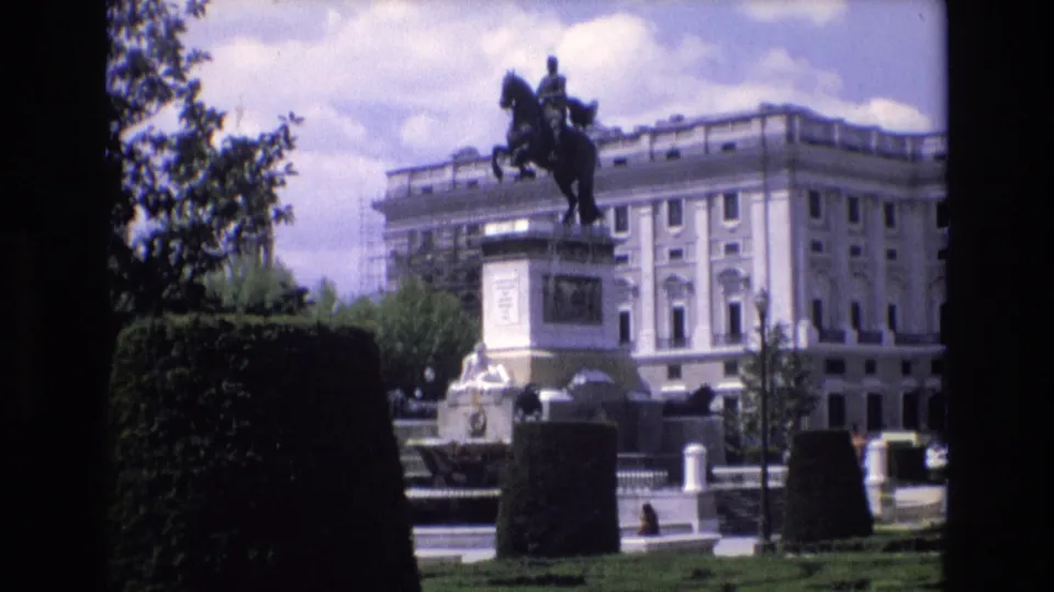 1972 Spain 8mm frame showing the Monument to Philip IV in Plaza de Oriente from across trimmed hedges, with a palatial facade and a few pedestrians in the background.