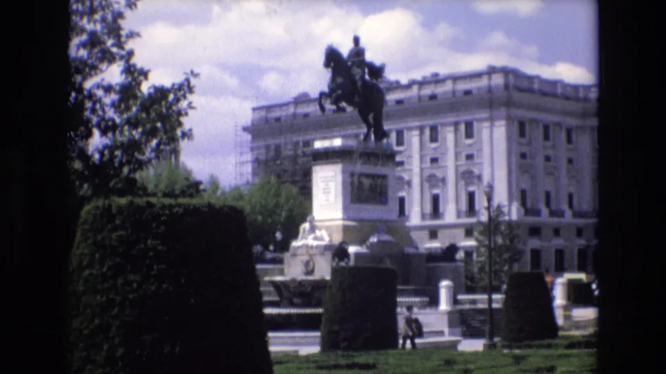 1972 Spain 8mm frame of the Plaza de Oriente monument, with the equestrian statue silhouetted against sky and a large palatial building while pedestrians move through the plaza.
