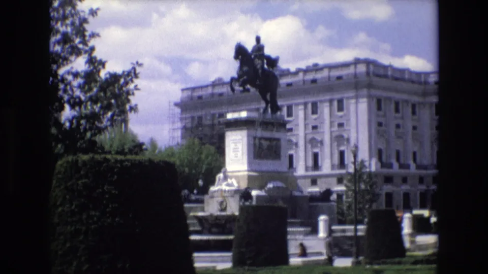 1972 Spain home-movie frame showing the Monument to Philip IV and Plaza de Oriente gardens, with soft-focus hedges and a palace-like building behind the statue.