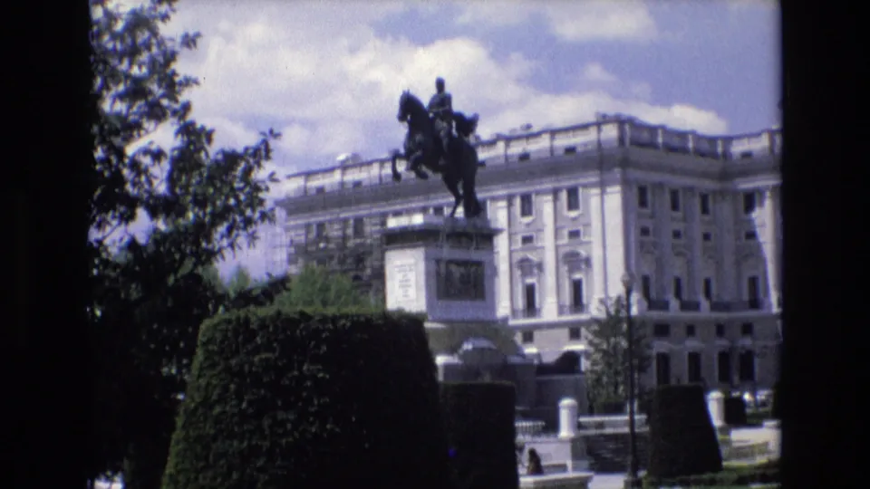 1972 Spain 8mm frame of the Monument to Philip IV in Plaza de Oriente, viewed from a slightly shifted handheld angle with gardens, pedestrians, and a palatial backdrop.