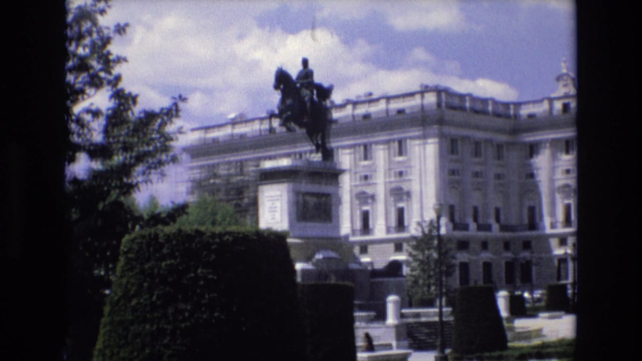 1972 Spain 8mm frame showing an equestrian statue in front of a grand pale palace facade, viewed from a shaded garden area with hedges and trees.