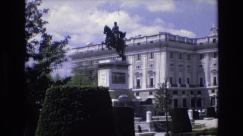1972 Spain 8mm frame showing an equestrian statue in front of a grand pale palace facade, viewed from a shaded garden area with hedges and trees.